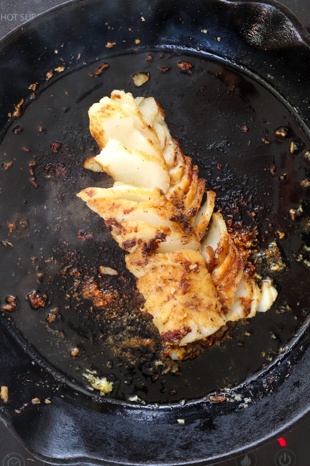 A zoomed in image of a black cast iron pan. There is fully browned garlic and cajun seasoing stuck to the pan. In the middle is pan-seared cod. The cod is starting to flake apart and the cajun coating is blackening.