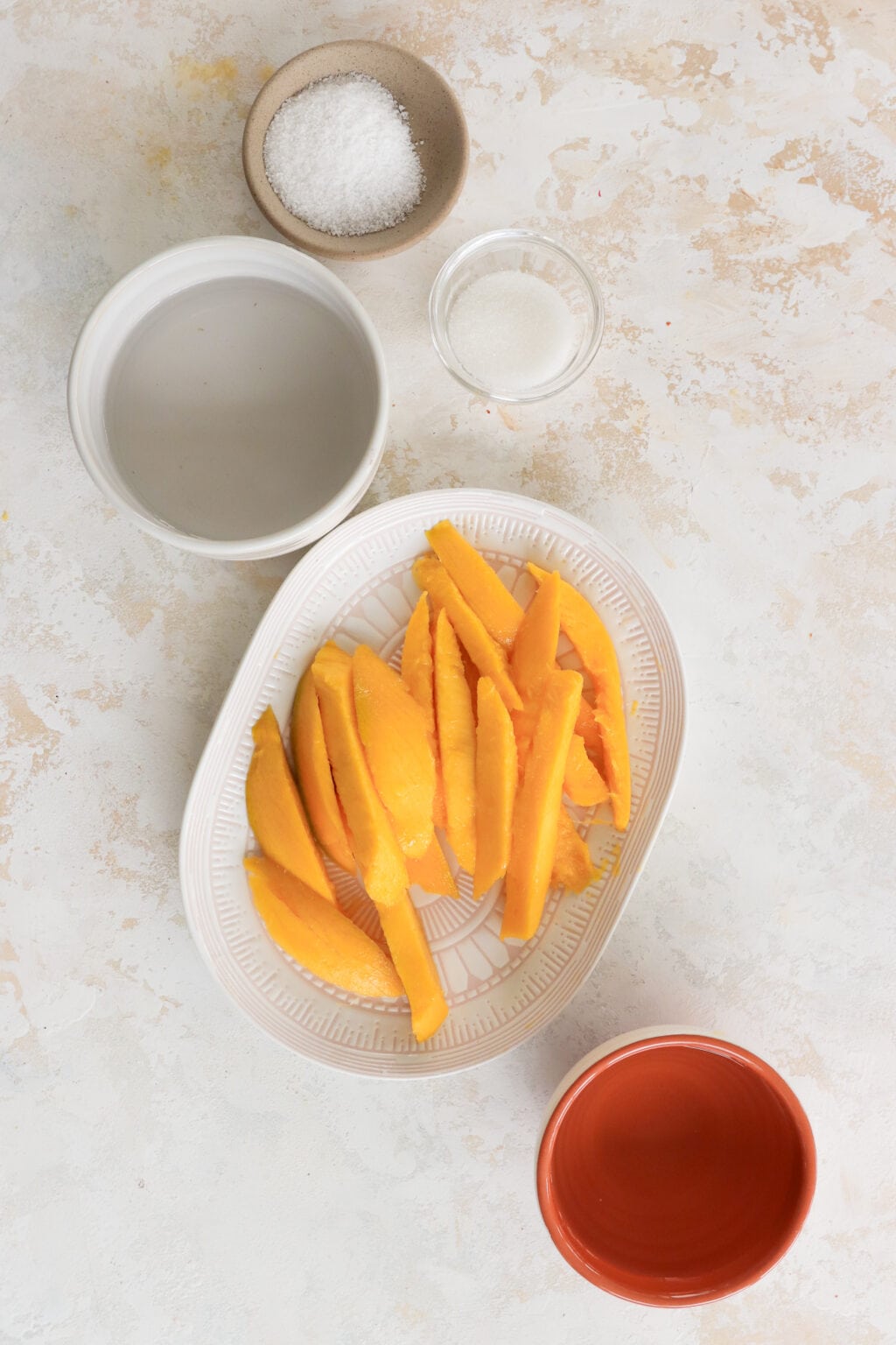 An overhead shot of the ingredients to make pickled mango. There is a bowl with sugar in it, a bowl with salt, a plate with sliced mango, and a bowl with white wine vinegar. 