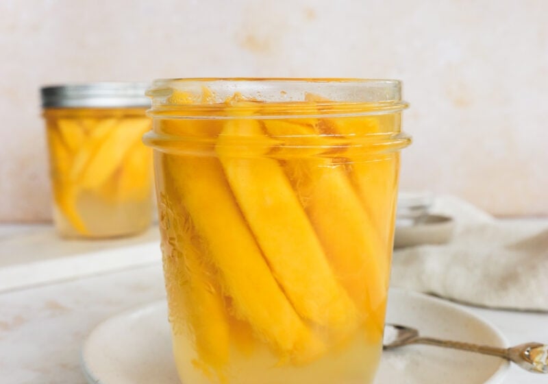 A mason jar filled with mangoes is on a white plate. There is a spoon to the right of the plate. In the background is another mason jar with pickled mangoes.