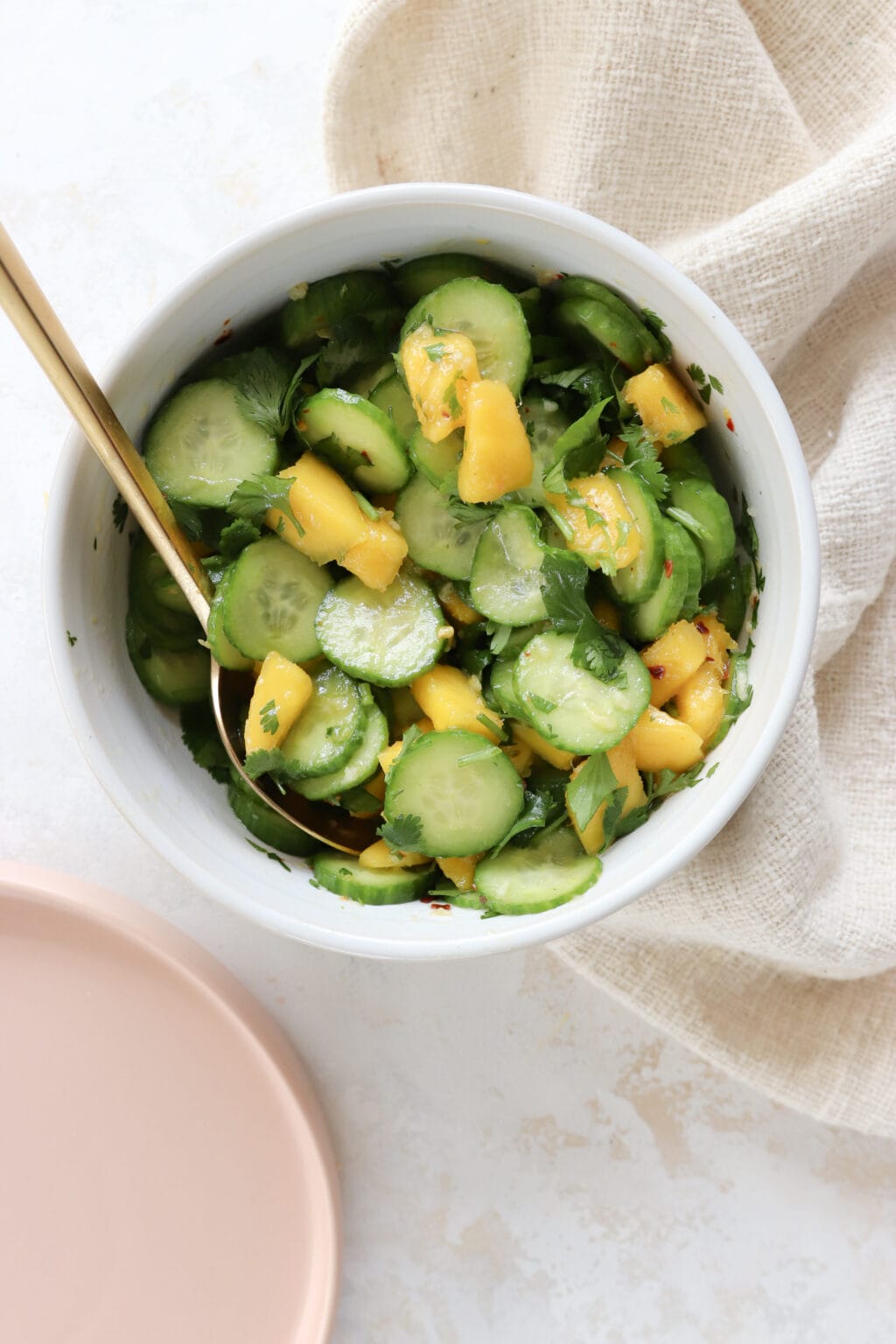 A white bowl is filled with a cucumber mango salad. There is a gold fork in it. To the left of the bowl is a pink plate. 