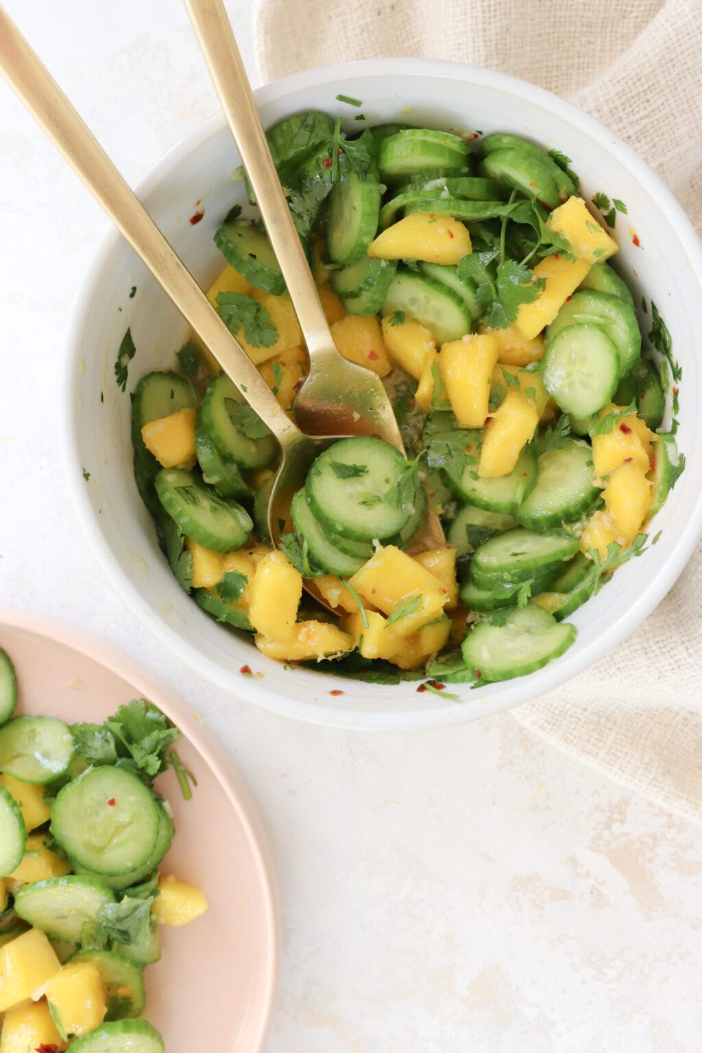In the left bottom corner is a pink plate. It has sliced cucumber and diced mango. A white bowl is in the middle it is filled with sliced cucumber and diced mango. There are two gold spoons serving the salad