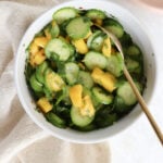 A white bowl is filled with sliced cucumber, diced mangoes, and cilantro. There is a gold spoon scooping out the salad