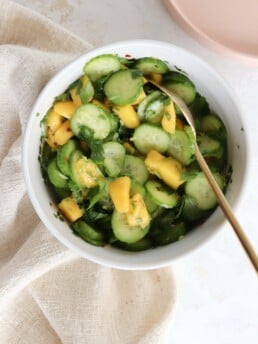 A white bowl is filled with sliced cucumber, diced mangoes, and cilantro. There is a gold spoon scooping out the salad