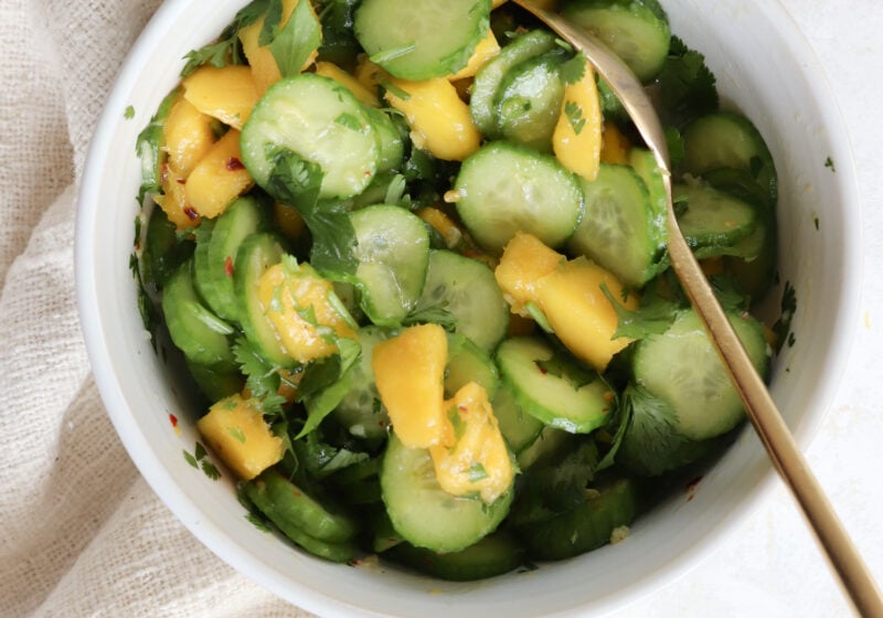 A white bowl is filled with sliced cucumber, diced mangoes, and cilantro. There is a gold spoon scooping out the salad