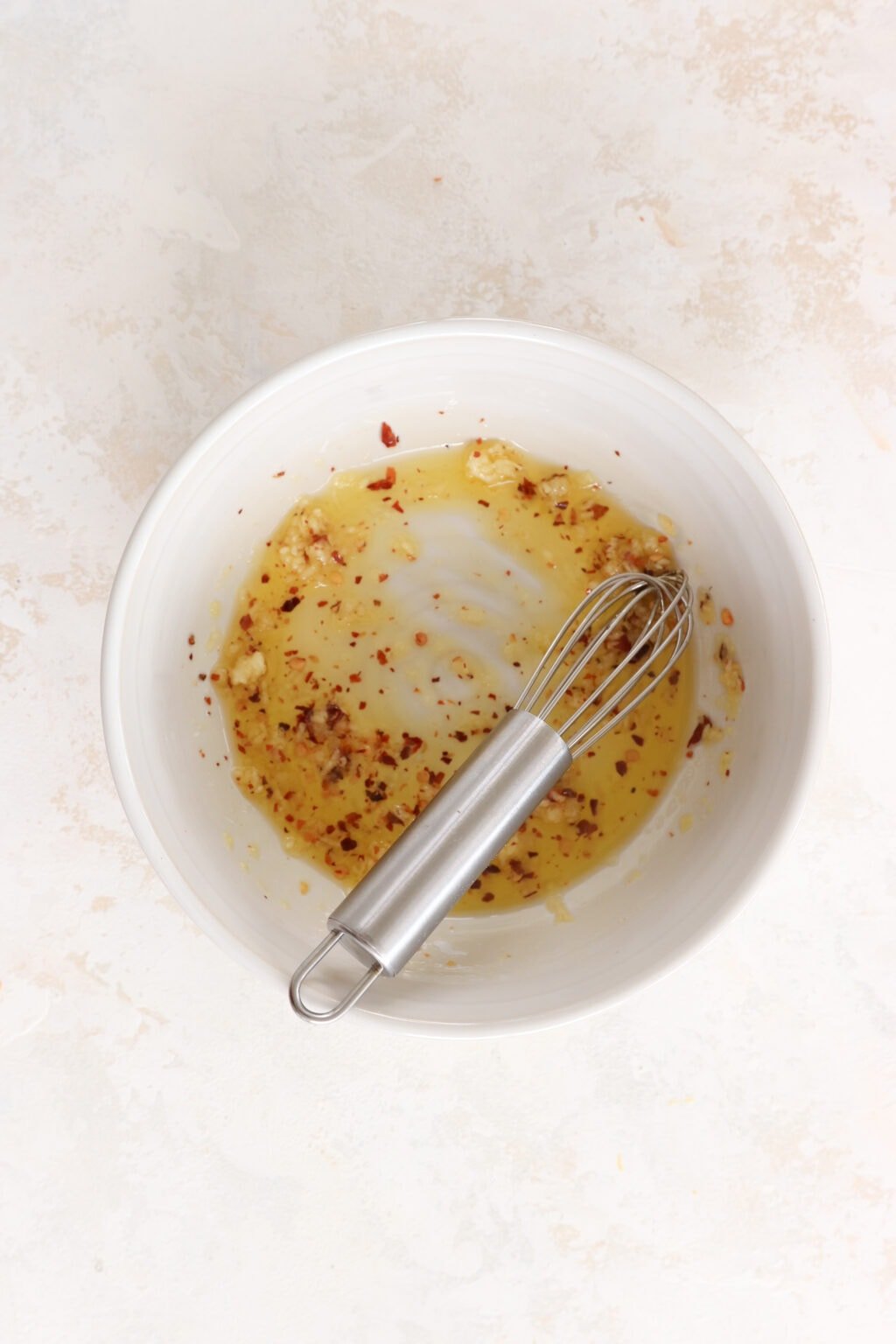 A small white bowl with a mini whisk is filled with oil, garlic, and chili flakes