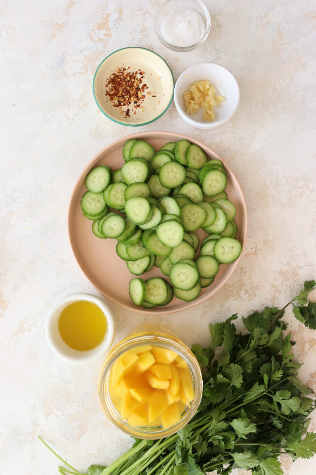 An overhead shot of the ingredients for the salad. At the top are two white bowls. One has red pepper flakes and the other has garlic. IN the middle there is a plate with sliced mini cucumber. At the bottom is a white bowl of olive oil, a jar of pickled mango, and a bunch of cilantro.