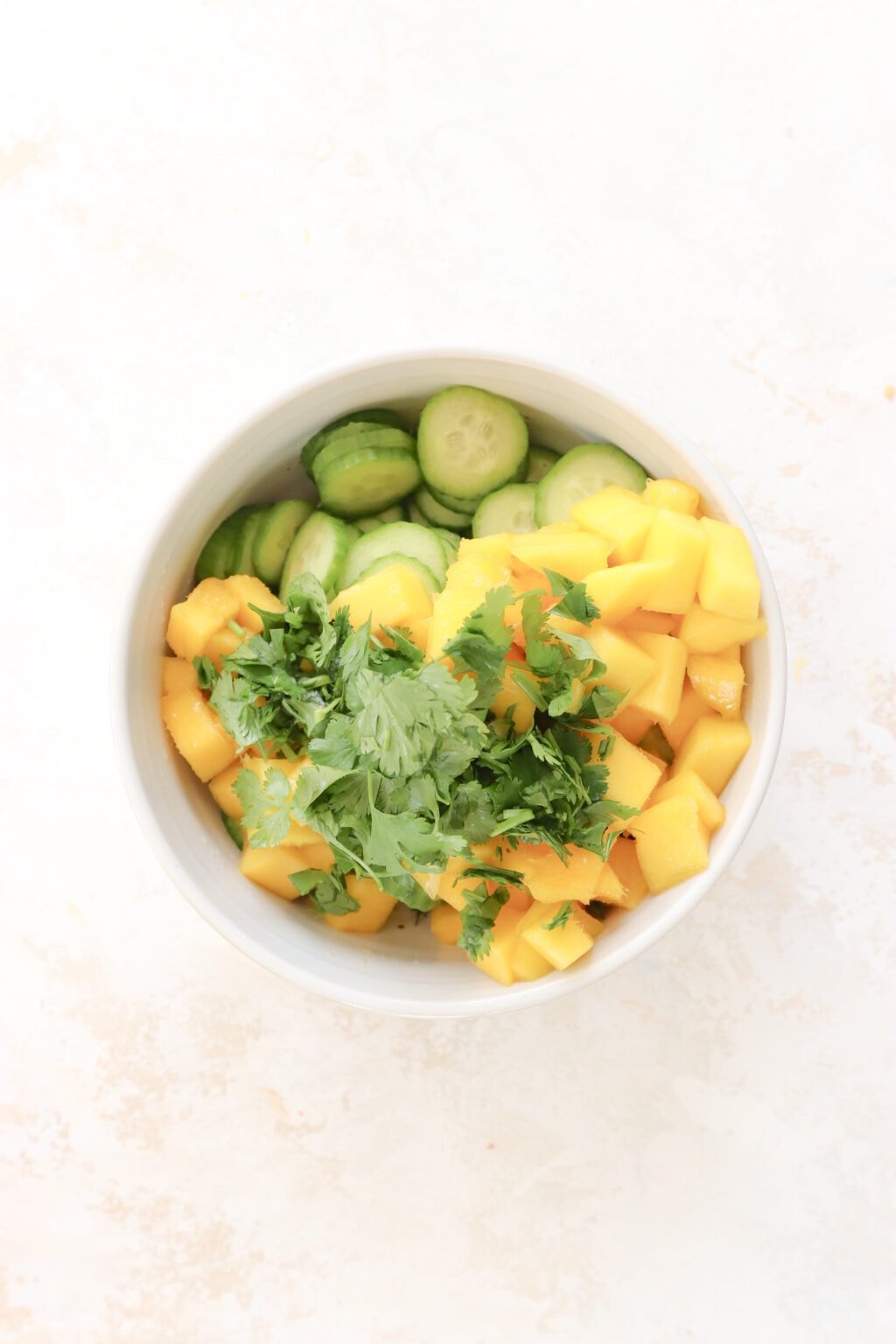 A white bowl is filled with sliced cucumber, diced mango, chopped cilantro