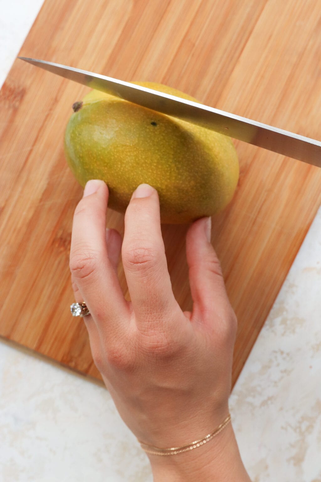 A mango that is green and yellow in color is on a  wooden cutting board. A chef's knife is cutting into theand a woman's hand is holding the mango. 