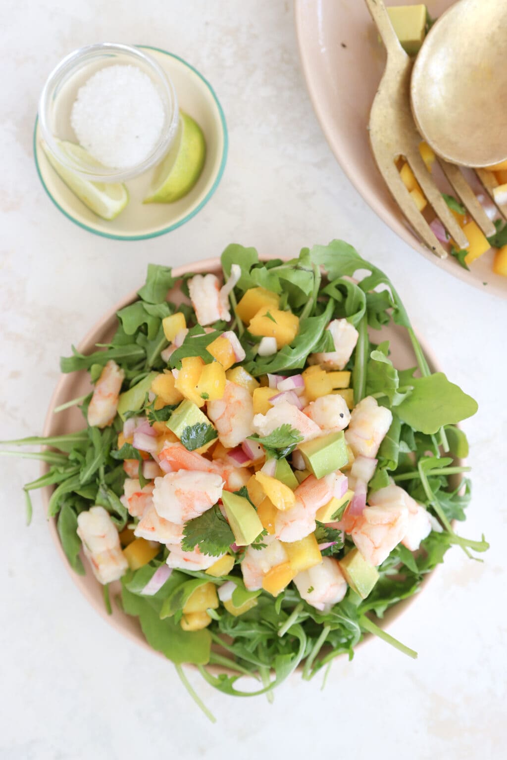 A pink plate with a heaping serving of salad that has arugula, avocado, mango, and shrimp. In the background is a platter of the same salad and two gold serving spoons.