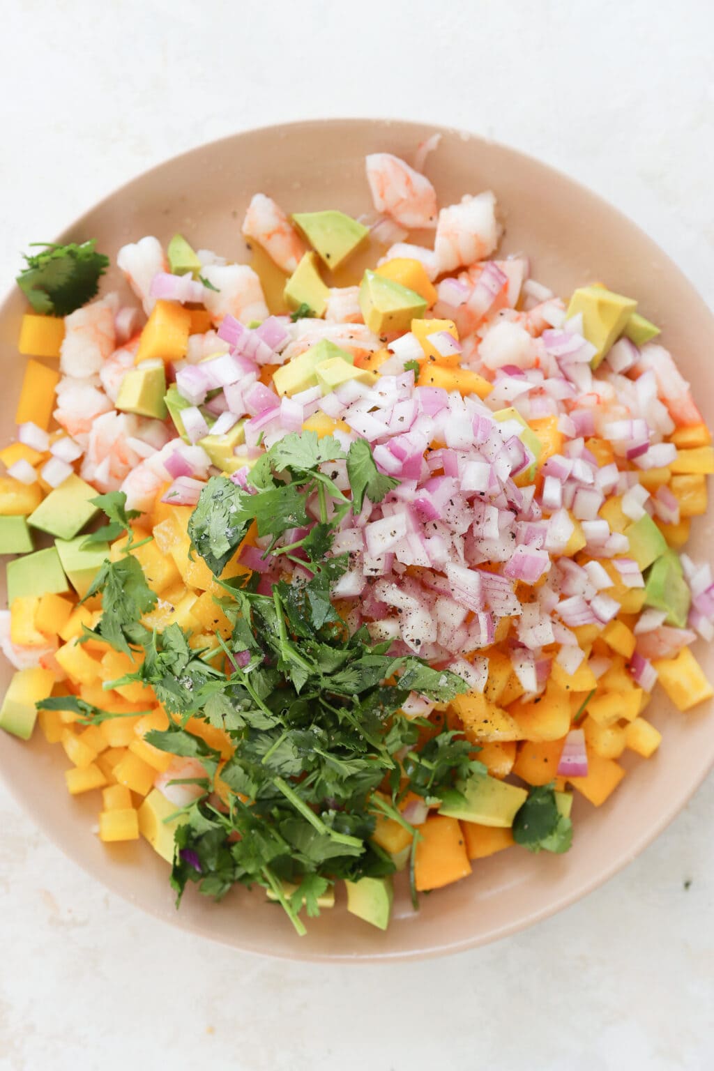 A pink plate with ingredients for a prawn ceviche, mango, avocado salad. The ingredients have not been mixed. There is cubed mango, chopped shrimp, cubed avocado, diced red onion, cilantro