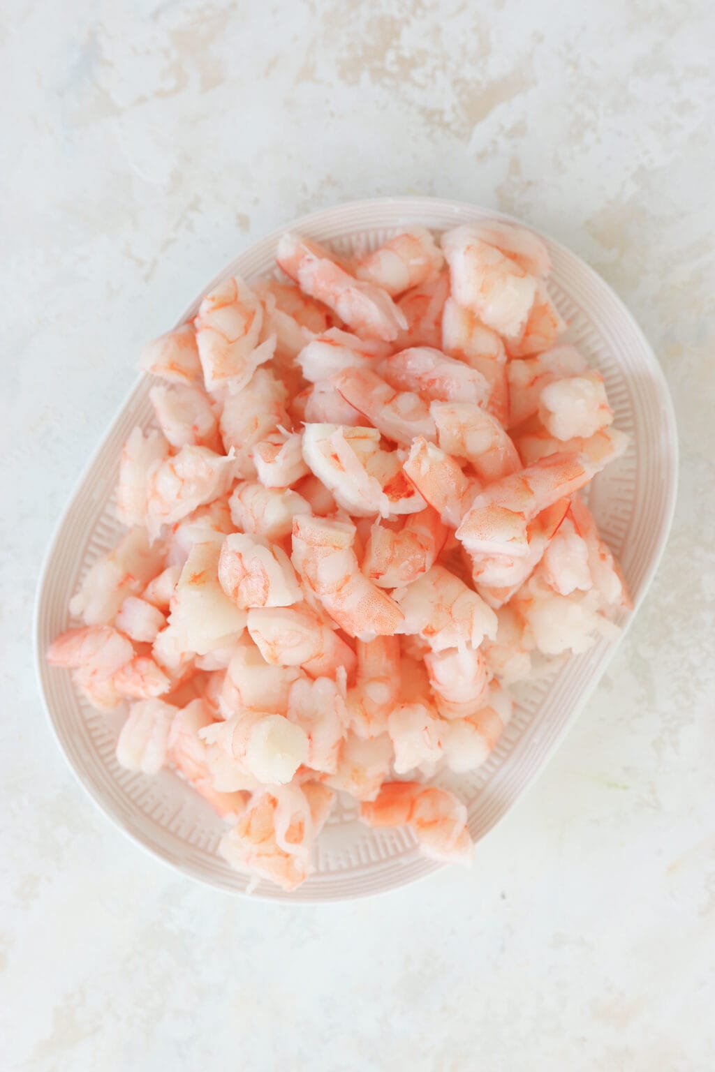 A white plate is on a marble background. There is pink shrimp piled on the plate. The shrimp is chopped into thirds