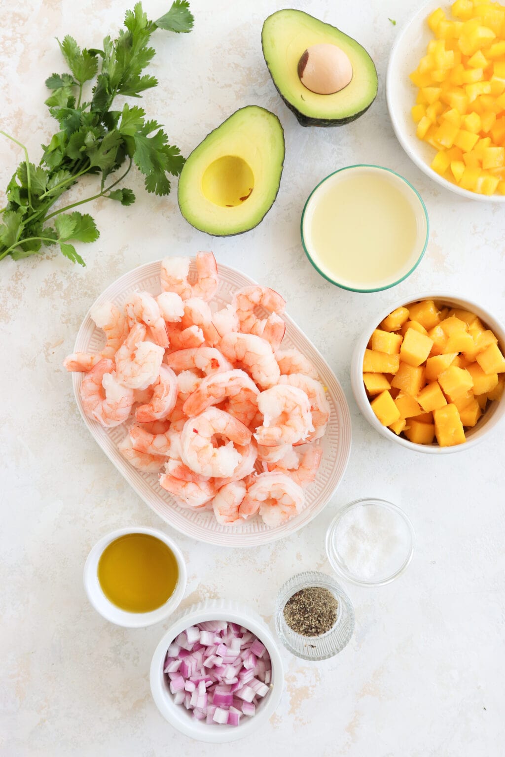 An overhead shot of the ingredients. From top to bottom left to right: A bundle of cilantro, half an avocado lime juice cubed yellow pepper. Next is pink shrimp and cubed mango. Bottom is olive oil, diced red onion, black pepper, and salt.