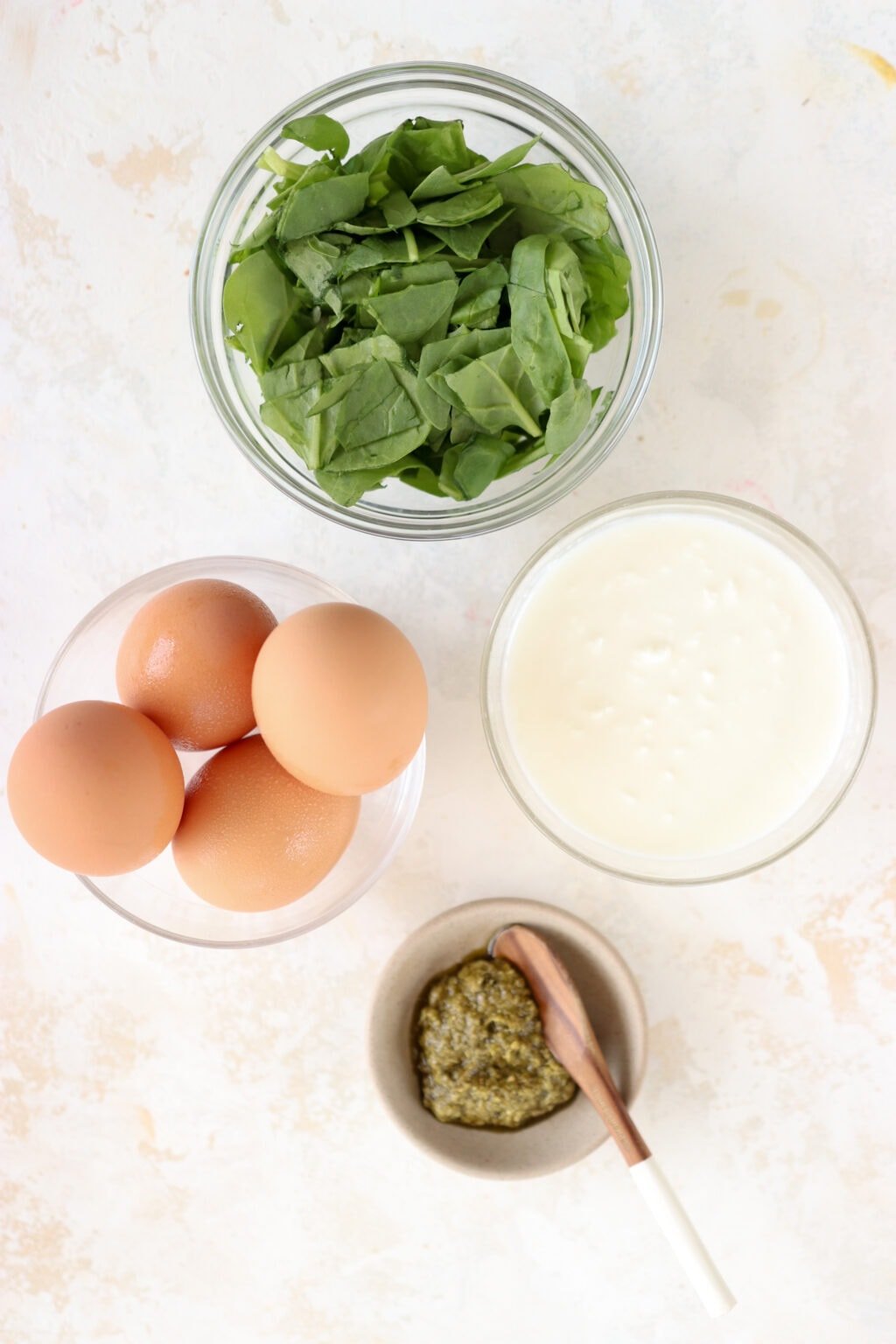The ingredients to make this recipe are laid out in bowls including spinach, cottage cheese, eggs, and pesto. 