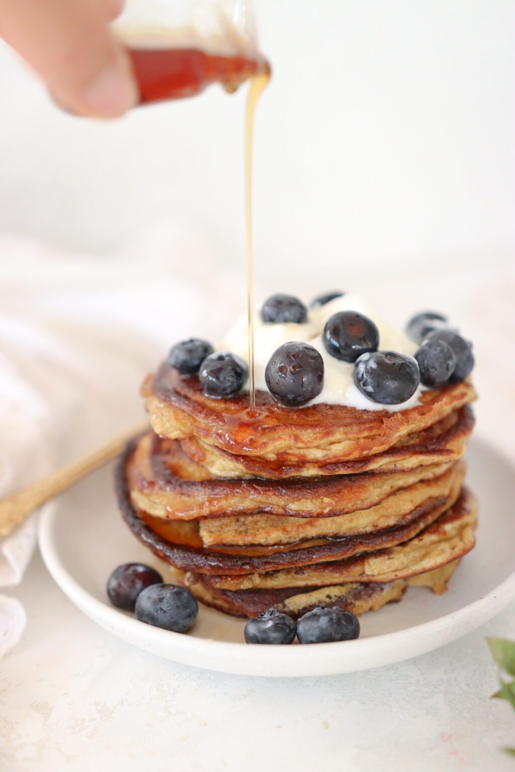 A stack of pancakes topped with yogurt and blueberries with a hand drizzling maple syrup over the stack.