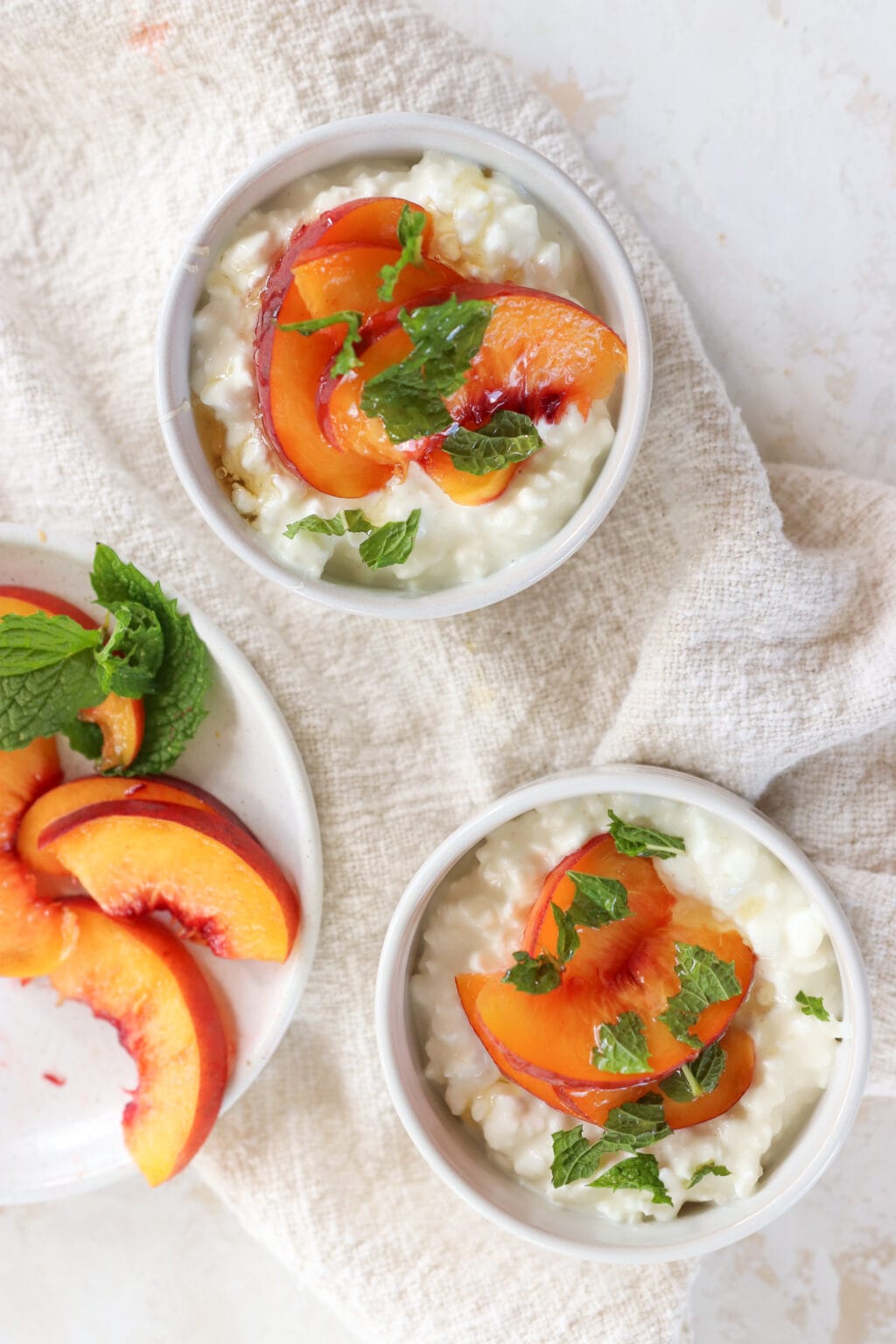 Two white bowls are filled with cottage cheese, sliced peaches and mint. To the left of the bowls is a plate of sliced peaches with mint 