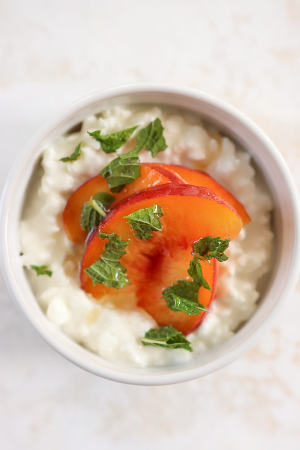 A white bowl is on an off white cloth. In the bowl is cottage cheese with slices of peaches topped with mint. There is a gold spoon beside the bowl.