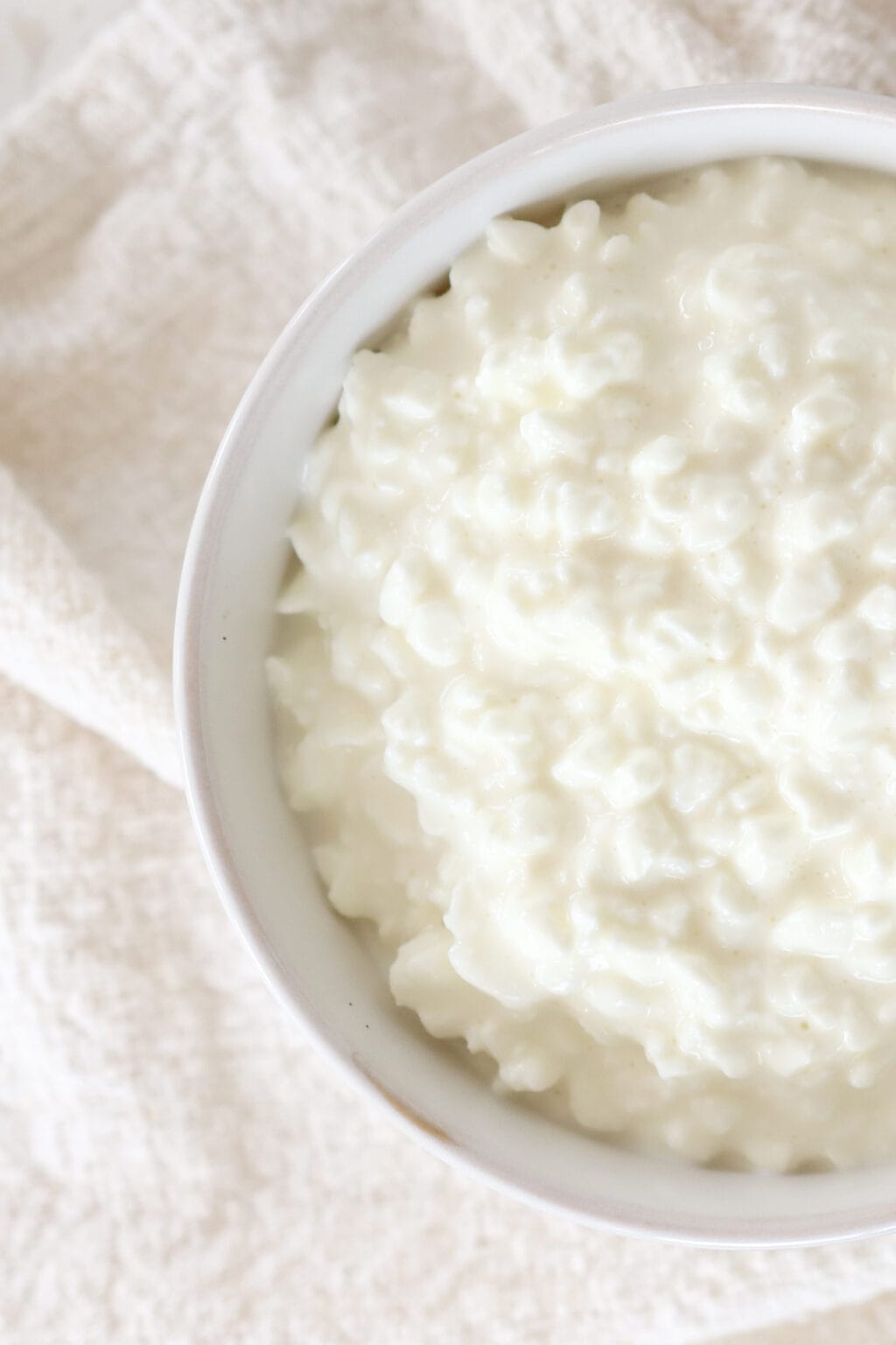 A zoomed in image of a white bowl with cottage cheese inside. The bowl is sitting on a textured dish towel.