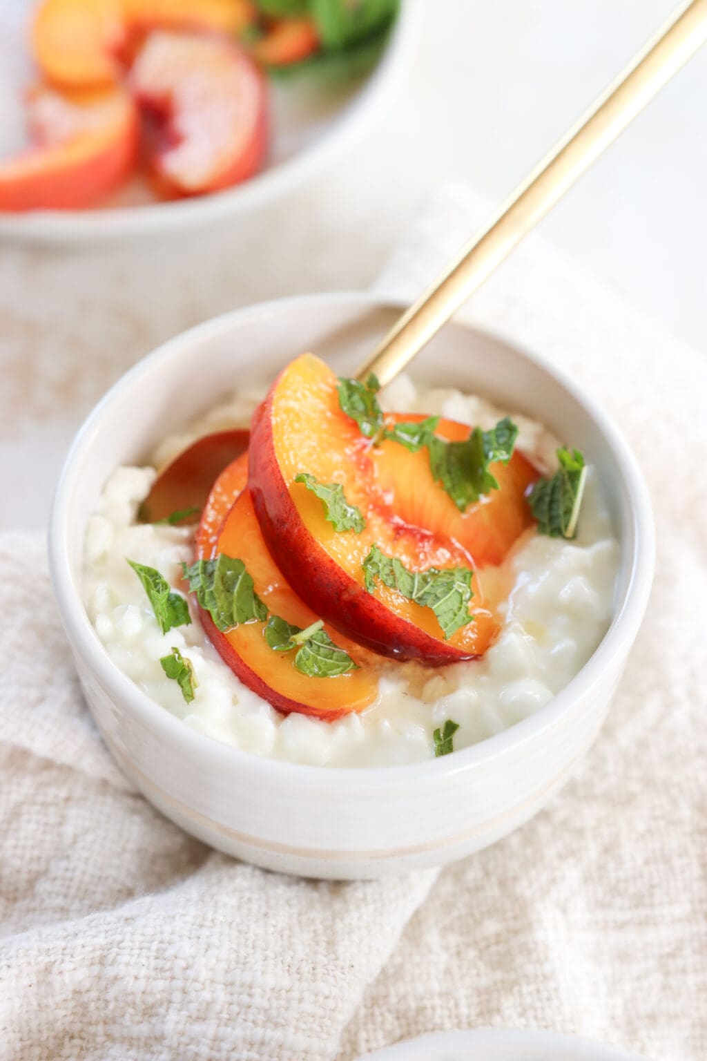 A white bowl is filled with cottage cheese, sliced peaches and topped with mint. There is a gold spoon in the bowl. 