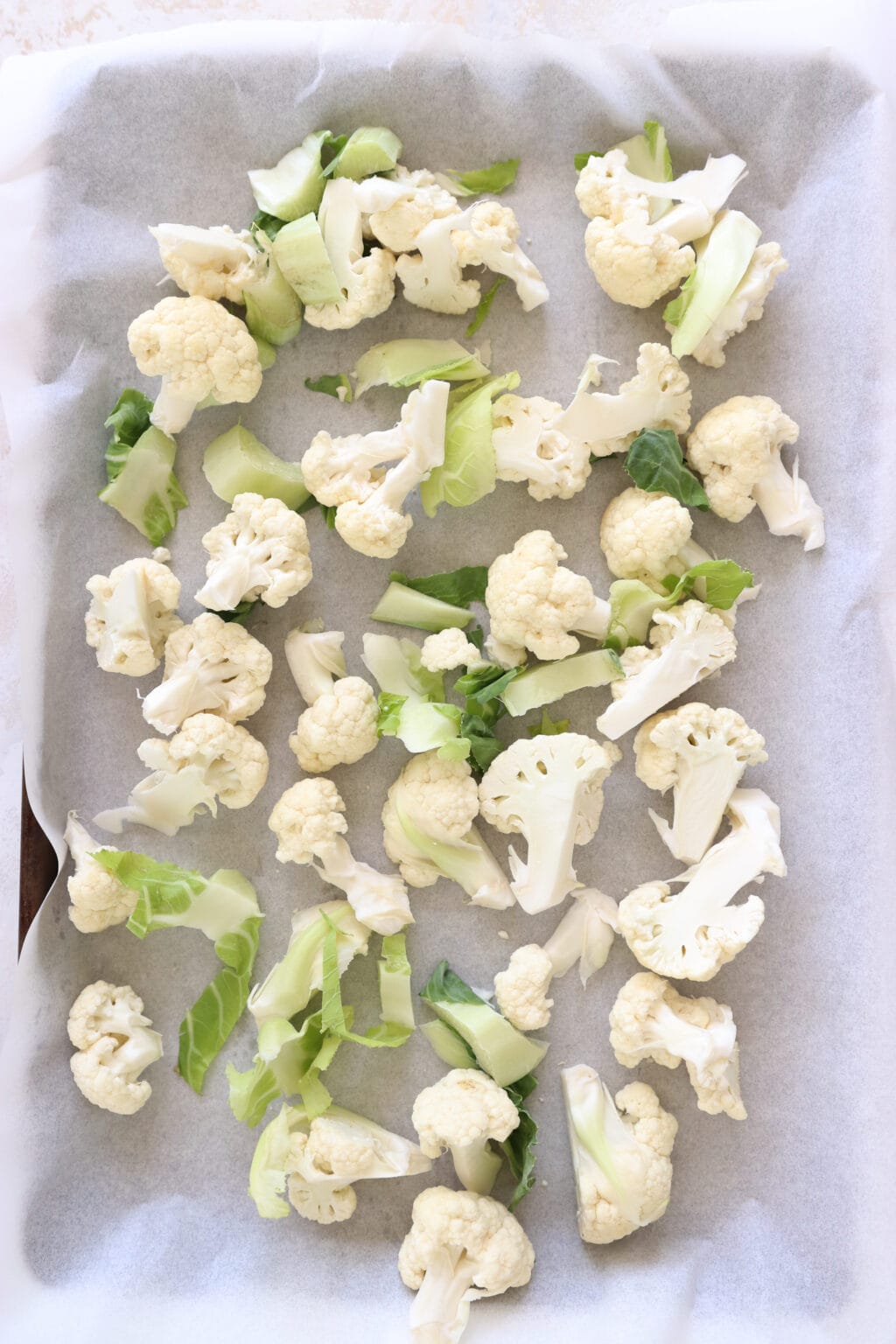 Raw cauliflower florets are on a parchment lined baking sheet. The cauliflower greens are also on the baking sheet.