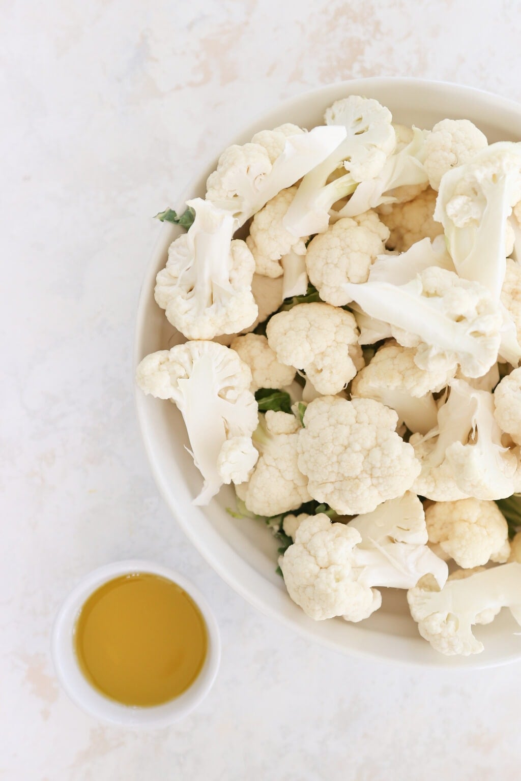 A white bowl holds cauliflower florets. Beside it is a white ramekin with brown oil in it.