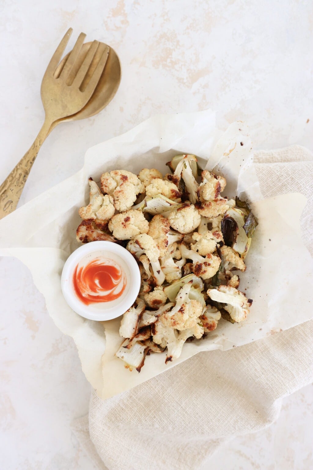 A bowl lined with parchment paper holds roasted cauliflower florets. There is a small ramekin filled with white yogurt and a swirl of hot sauce. There is a set of gold serving spoons beside the cauliflower bowl. 