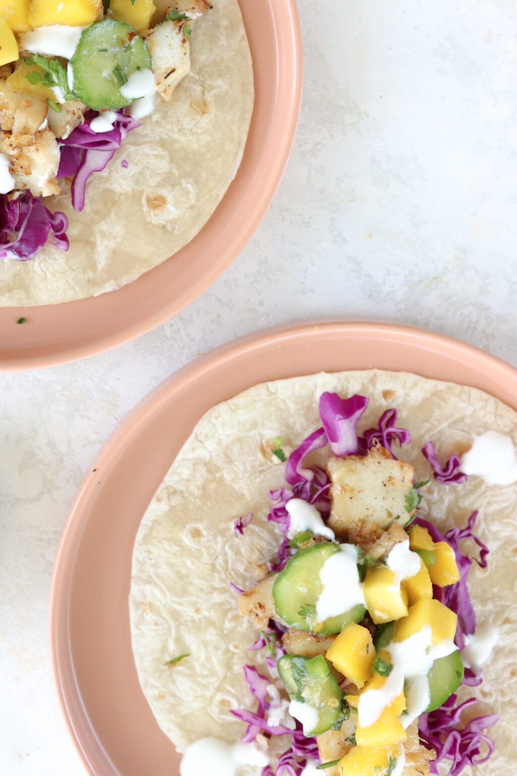 2 fish tacos with cabbage, mango, and cucumber filling are on pink plates.