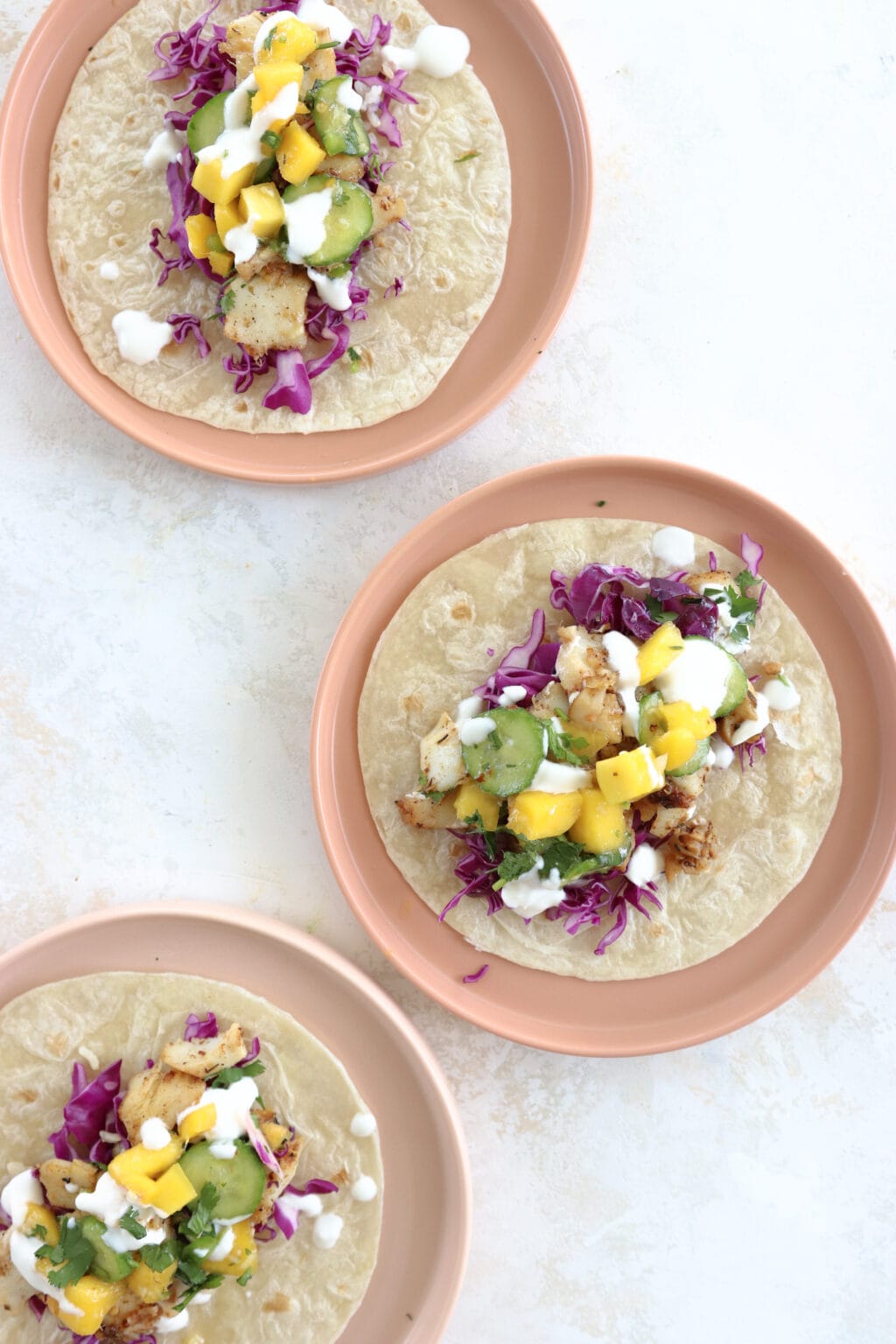 3 blackened fish tacos topped with cabbage slaw, mango and cucumber on pink plates.