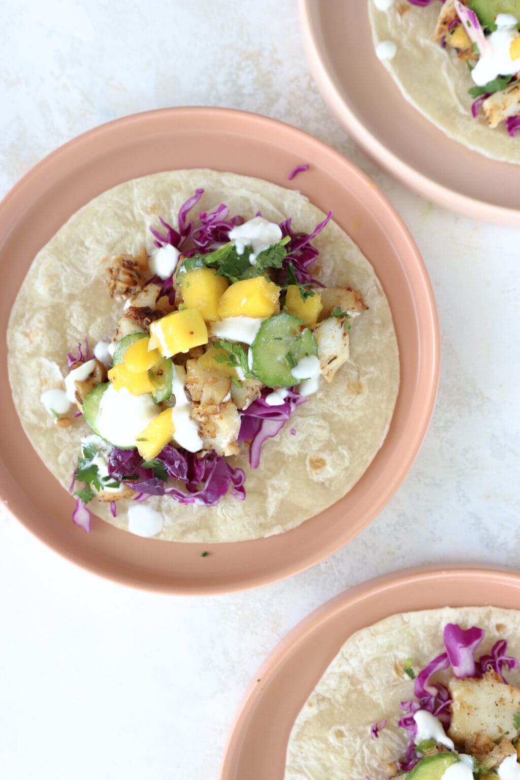 A fish taco with cabbage, mango, and cucumber toppings is on a plate.