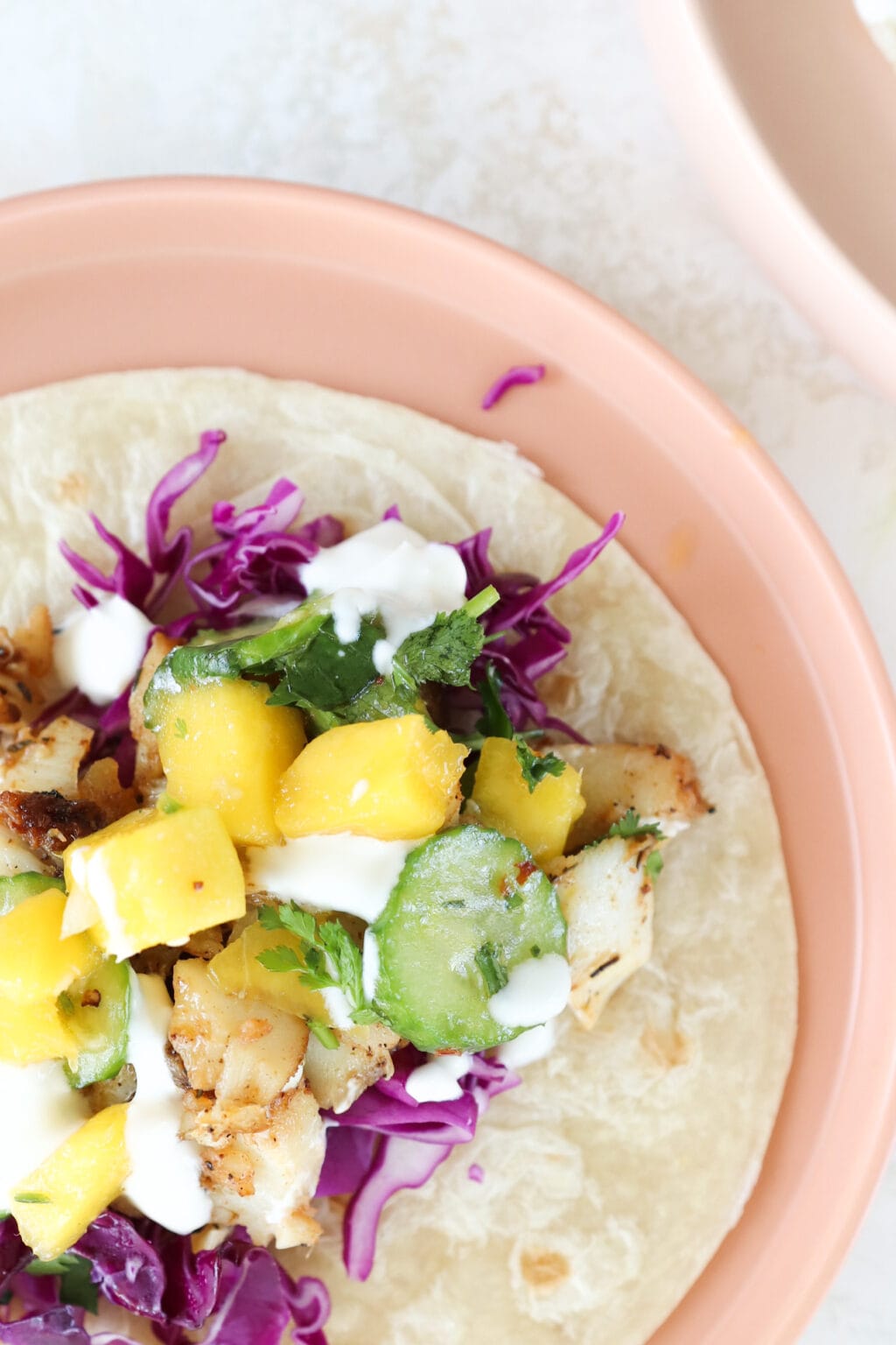 A fish taco with cabbage, cucumber, and mango as filling is on a plate.