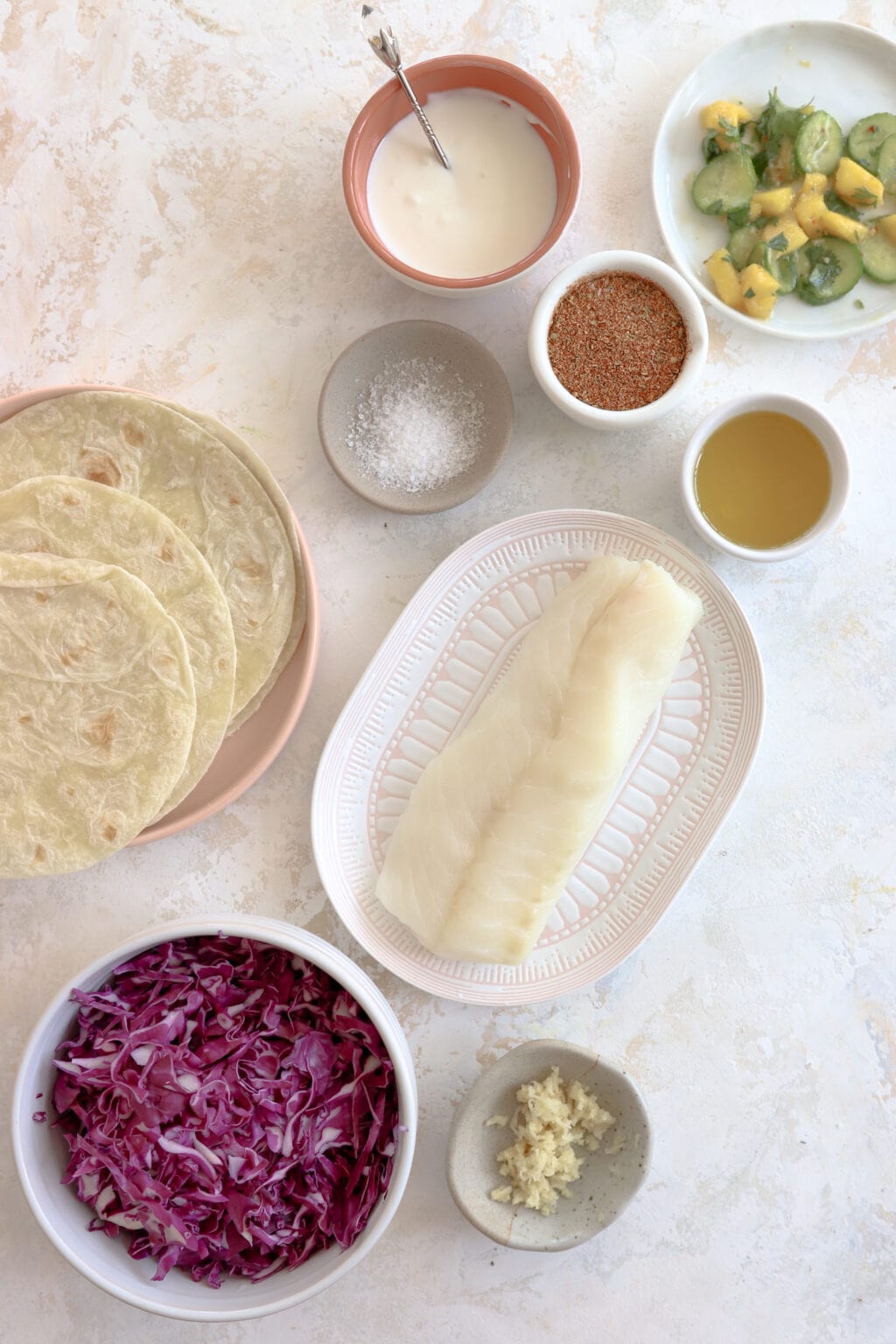 Ingredients to make fish tacos placed in bowls and on plates. Ingredients include mango and cucumber salad, lime crema, cajun spice, oil, salt, tortillas, white fish, shredded cabbage, and garlic.