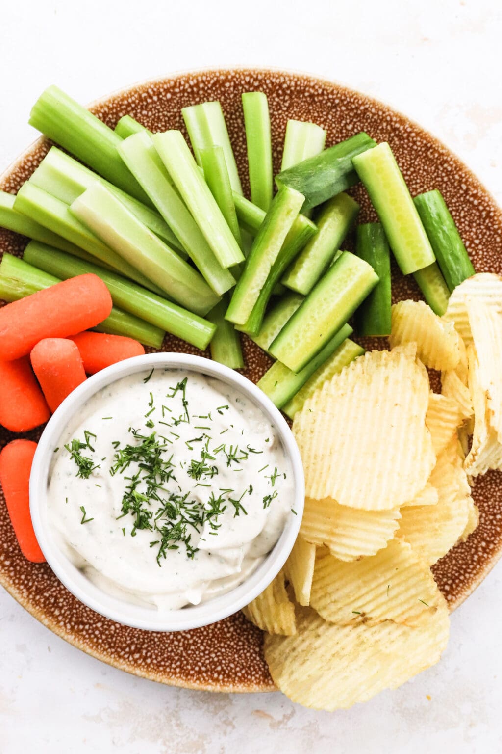Celery, carrots, and ripple chips on a brown plate with a white bowl of french onion cottage cheese dip