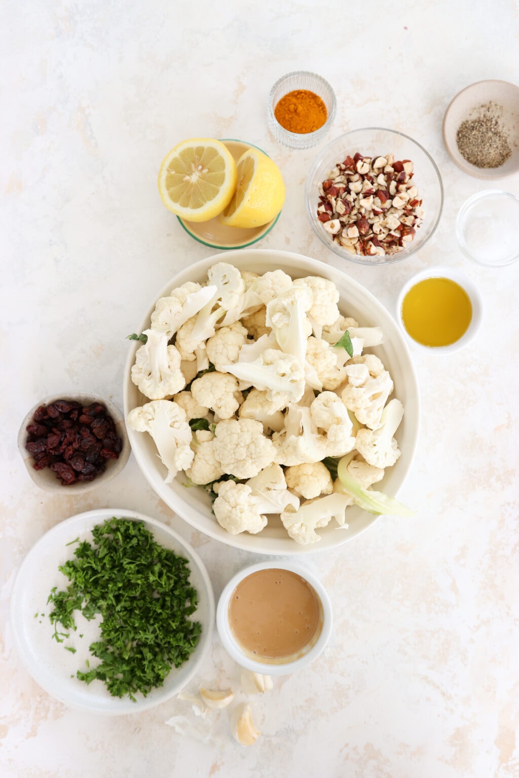 Ingredients for Curry Roasted Cauliflower with Lemon Tahini Sauce in white bowls. Ingredients include cauliflower florets, avocado oil, salt and pepper, curry powder, raisins, hazelnuts, parsley, tahini, lemon juice, olive oil, and garlic cloves