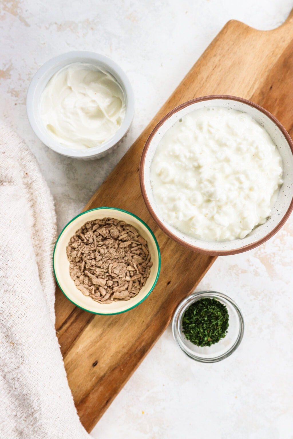 Small bowls holding ingredients for a french onion cottage cheese dip. Ingredients include cottage cheese, sour cream, french onion soup mix, and dried parsley