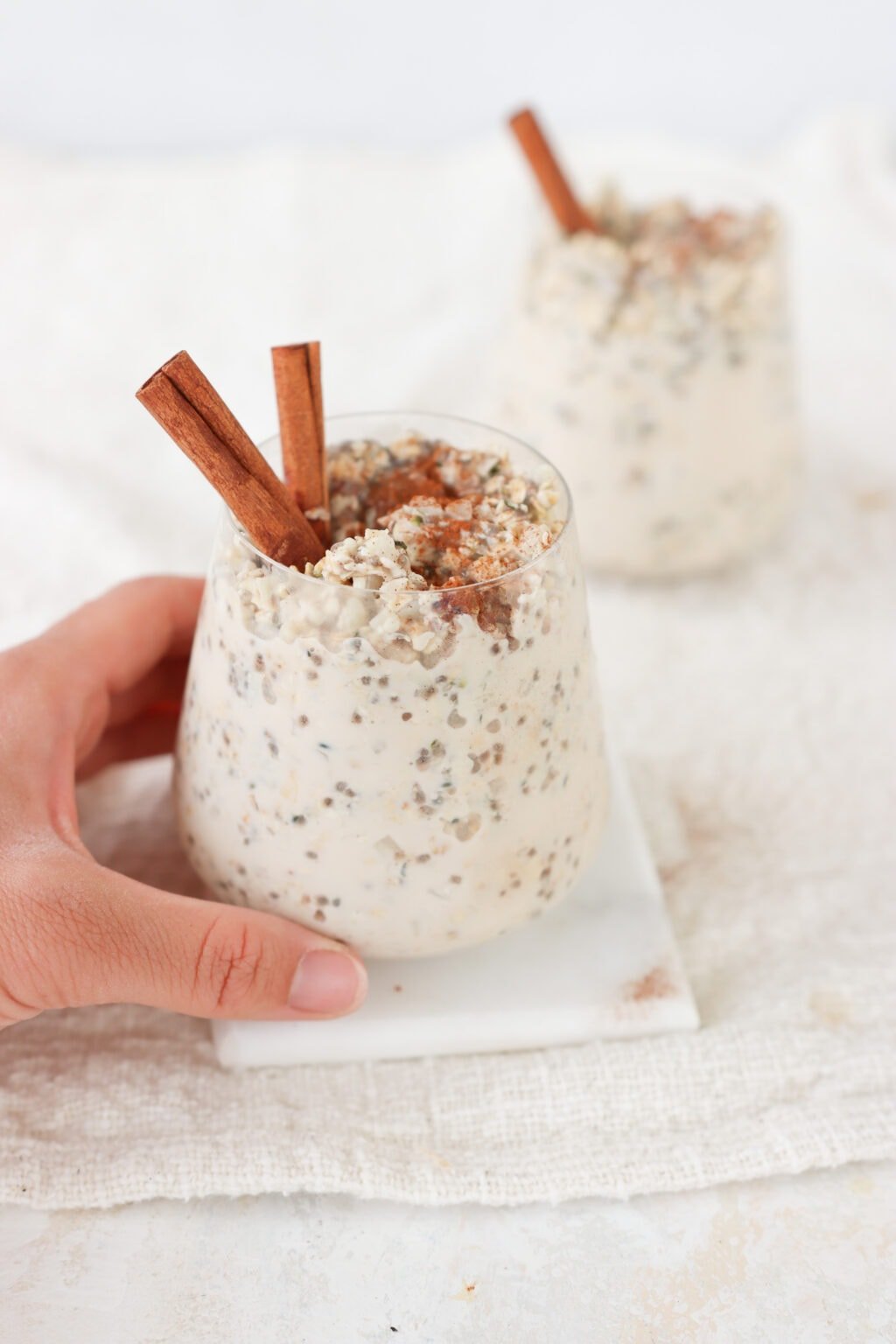 Two glasses on a white surface filled with meal prep cinnamon roll overnight oats with two cinnamon sticks and a hand picking up one of the glasses