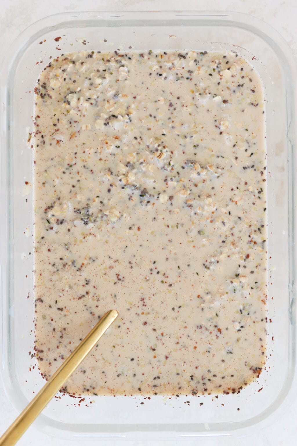 A glass container filled with mixed ingredients for cinnamon roll overnight oats, including quick oats, hemp hearts, chia seeds, vanilla extract, milk, maple syrup, salt, ground cinnamon, and cauliflower rice