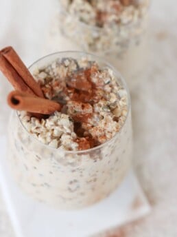 Two glasses on a white surface filled with meal prep cinnamon roll overnight oats with two cinnamon sticks