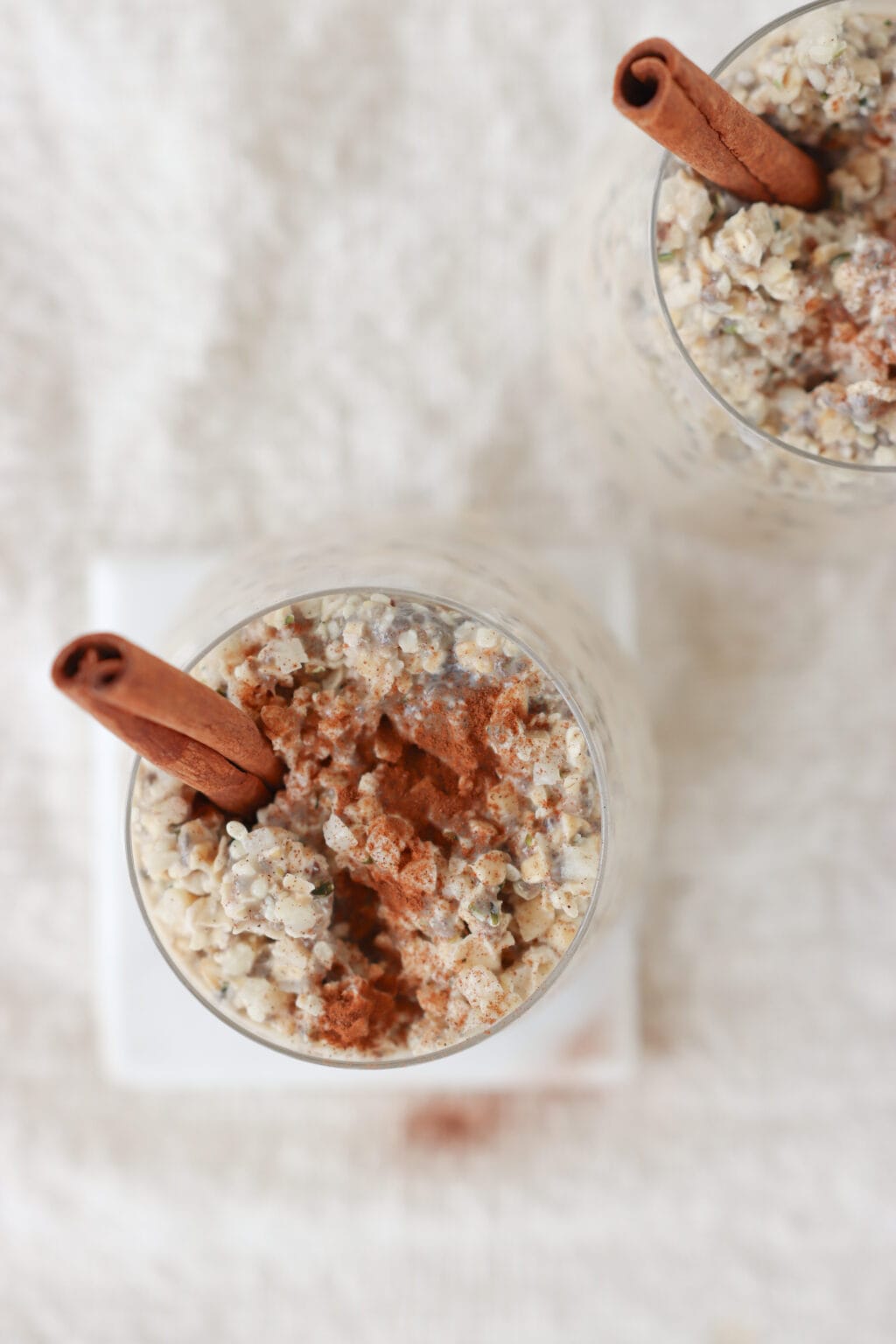 Two glasses on a white surface filled with meal prep cinnamon roll overnight oats with two cinnamon sticks