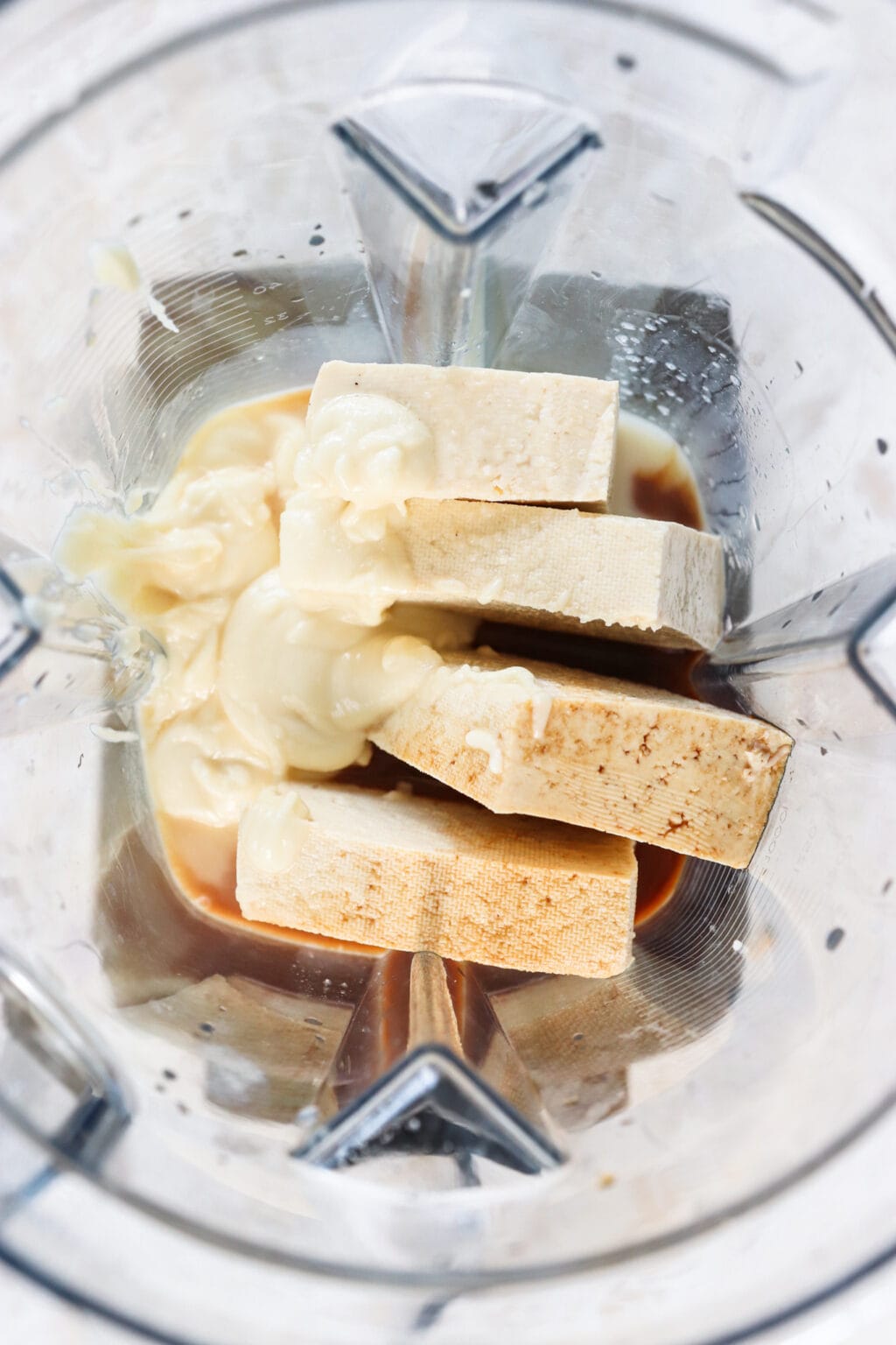 Ingredients for a 5 ingredient tofu eggnog mini creamy cheesecake, including tofu, white chocolate, eggnog, vanilla, coconut oil, and salt in a blender