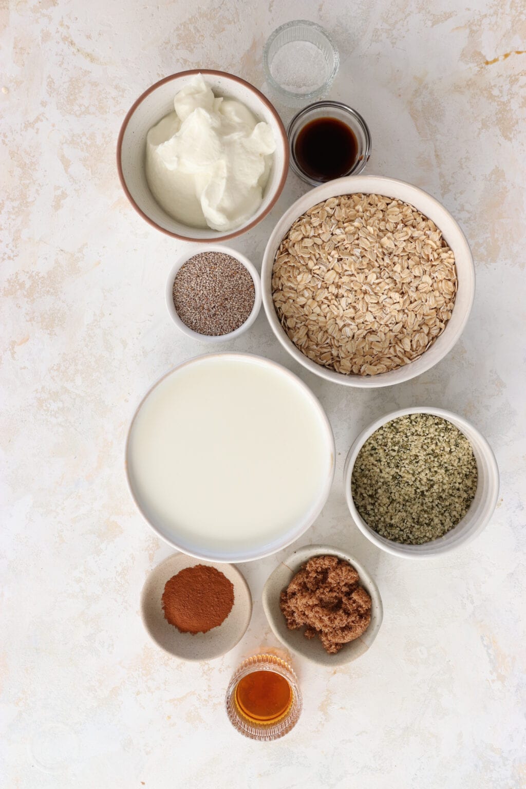 Ingredients for brown sugar and Greek yogurt overnight oats in white glass bowls, including quick oats, hemp hearts, chia seeds, vanilla extract, milk, cinnamon, brown sugar, maple syrup, salt, and Greek yogurt