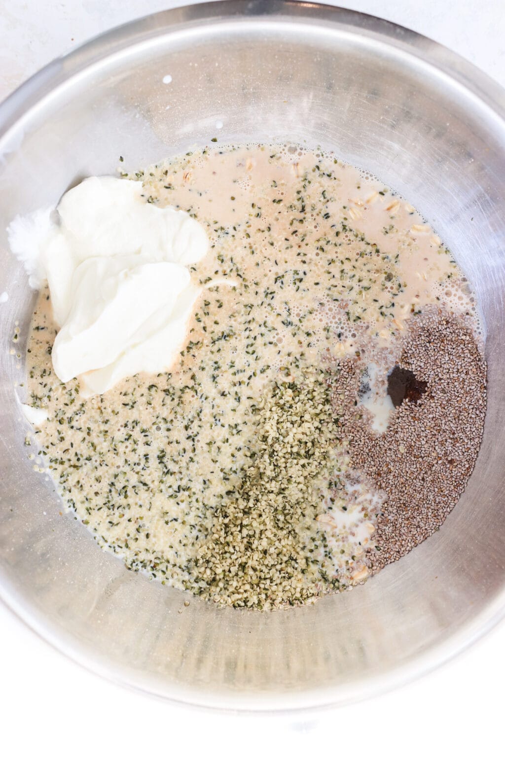 Ingredients for brown sugar and Greek yogurt overnight oats in a large bowl, including quick oats, hemp hearts, chia seeds, vanilla extract, milk, cinnamon, brown sugar, maple syrup, salt, and Greek yogurt