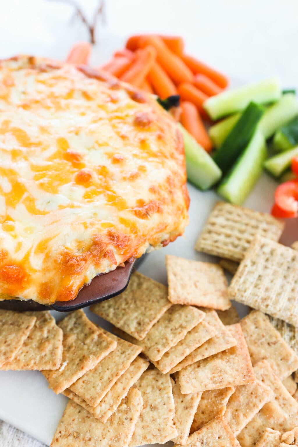 A dish of healthy white bean, spinach and artichoke dip on a white platter with crackers, cucumbers, and carrots for dipping