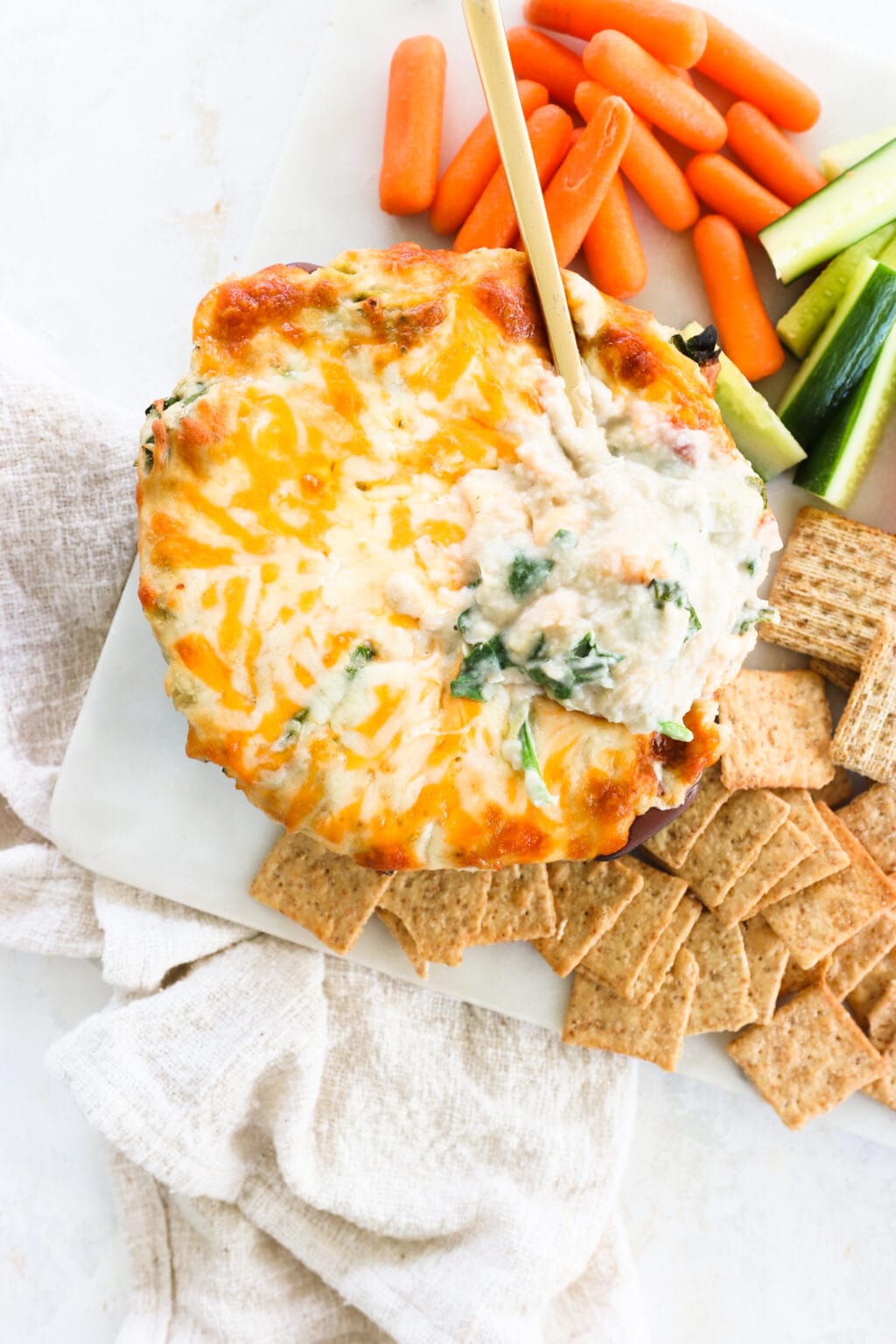 A dish of healthy white bean, spinach and artichoke dip on a white platter with crackers, cucumbers, and carrots for dipping