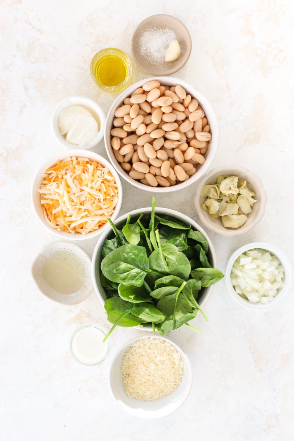 Ingredients for healthy white bean, spinach and artichoke dip, including white cannellini beans, plain Greek yogurt, olive oil, milk, garlic, salt, lemon juice, white onion, parmesan cheese, artichoke hearts, spinach, and marbled cheddar cheese