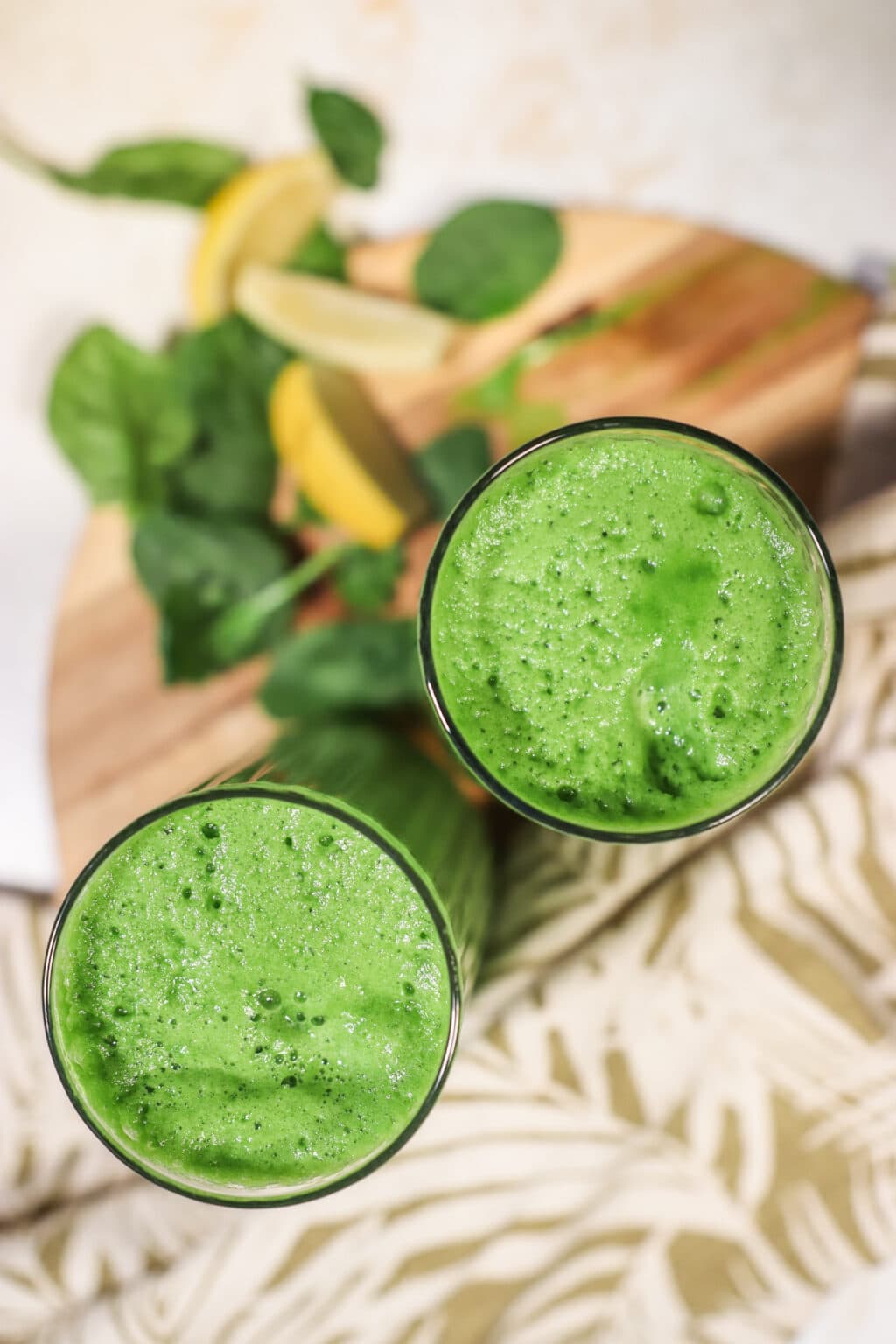 Two glasses filled with Blender Juice With Spinach, Pineapple & Cucumber, on a wood cutting board with lemon slices and spinach leaves on the side.