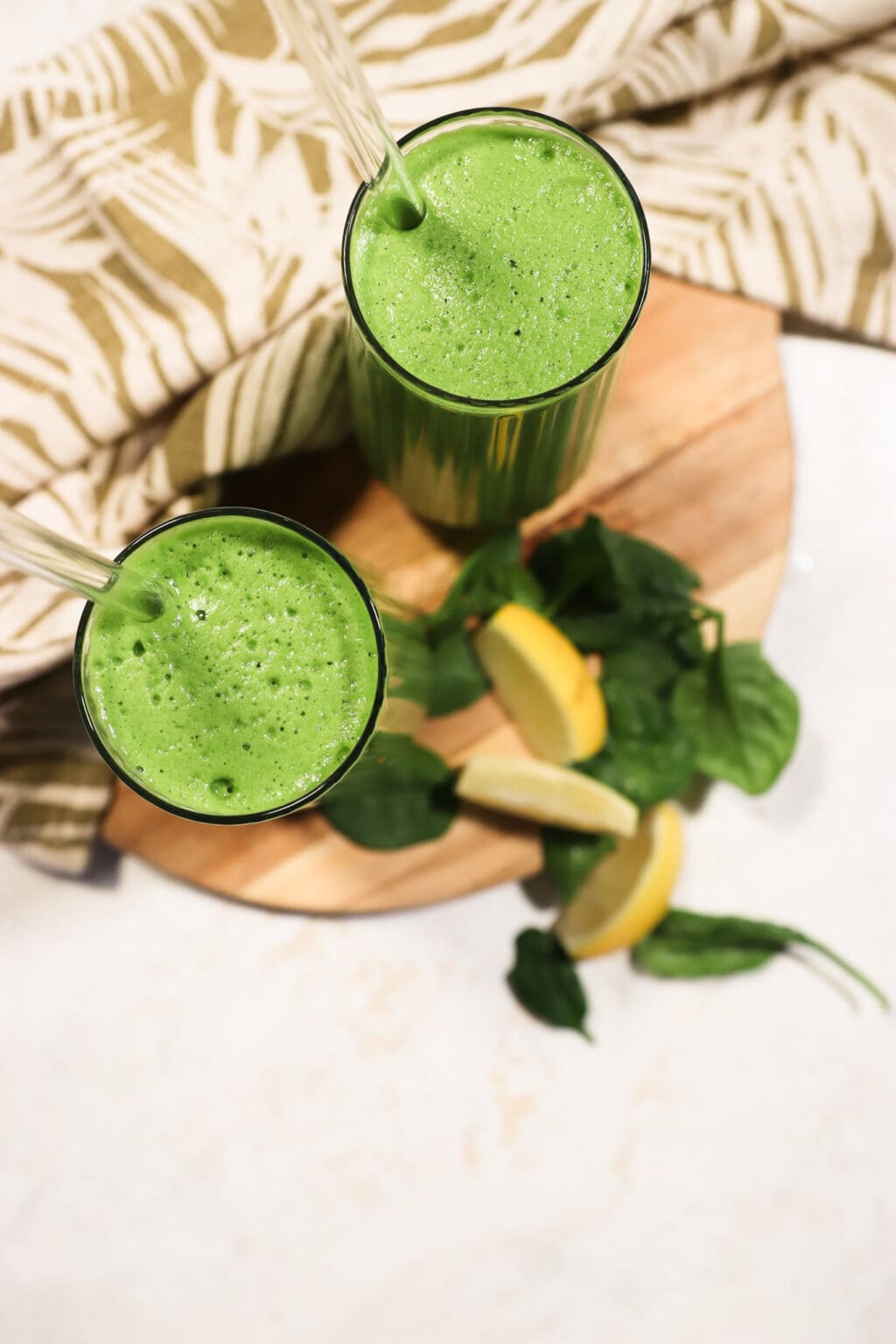 Two glasses filled with Blender Juice With Spinach, Pineapple & Cucumber, on a wood cutting board with lemon slices and spinach leaves on the side.