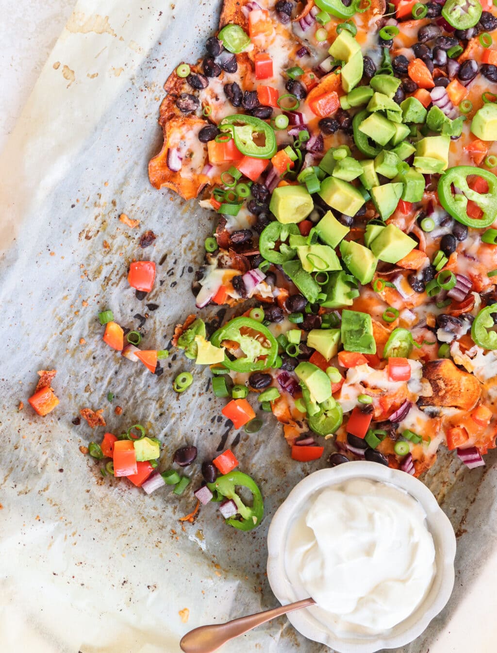 Partially eaten Healthy Black Bean & Bell Pepper Sweet Potato Nachos on a baking sheet with parchment paper and a small bowl of sour cream