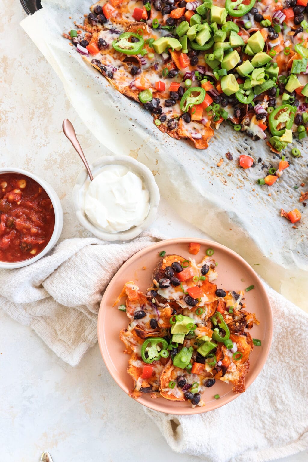 Healthy Black Bean & Bell Pepper Sweet Potato Nachos on a pink plate, with sour cream and salsa in small bowls on the side, and the baking sheet of cooked nachos at the top