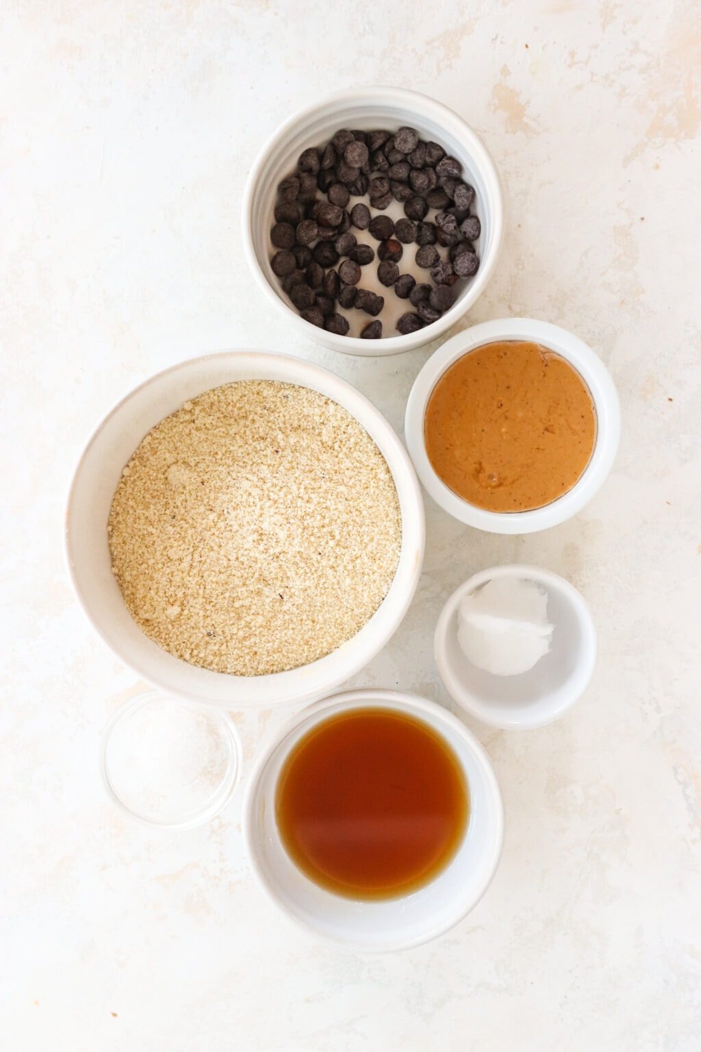 Ingredients for No Bake Peanut Butter & Chocolate Protein Bars in white glass bowls, including almond flour, peanut butter, maple syrup, salt, coconut oil, dark chocolate, hemp hearts or flaky salt