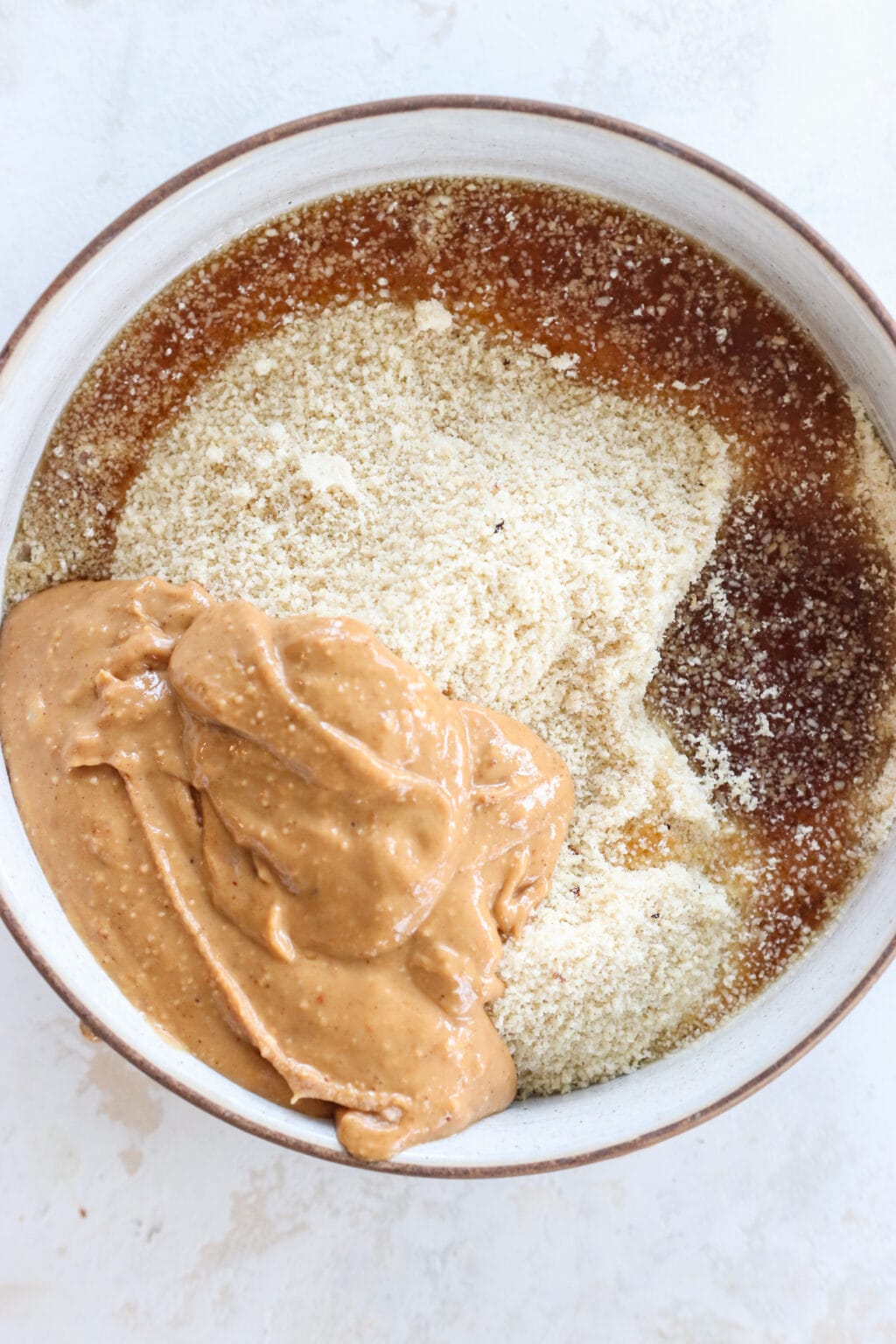 Ingredients for No Bake Peanut Butter & Chocolate Protein Bars in a white glass bowl, including almond flour, peanut butter, maple syrup, salt, coconut oil, dark chocolate, hemp hearts or flaky salt