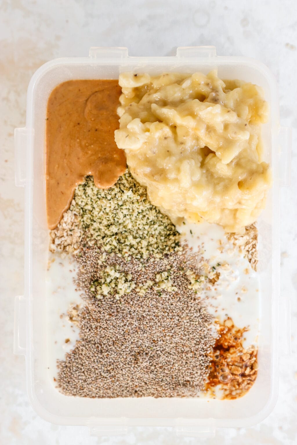 Ingredients for Peanut Butter & Banana Kefir Overnight Oats in a large container, including quick oats, hemp hearts, chia seeds, vanilla extract, plain kefir, maple syrup, peanut butter, and bananas